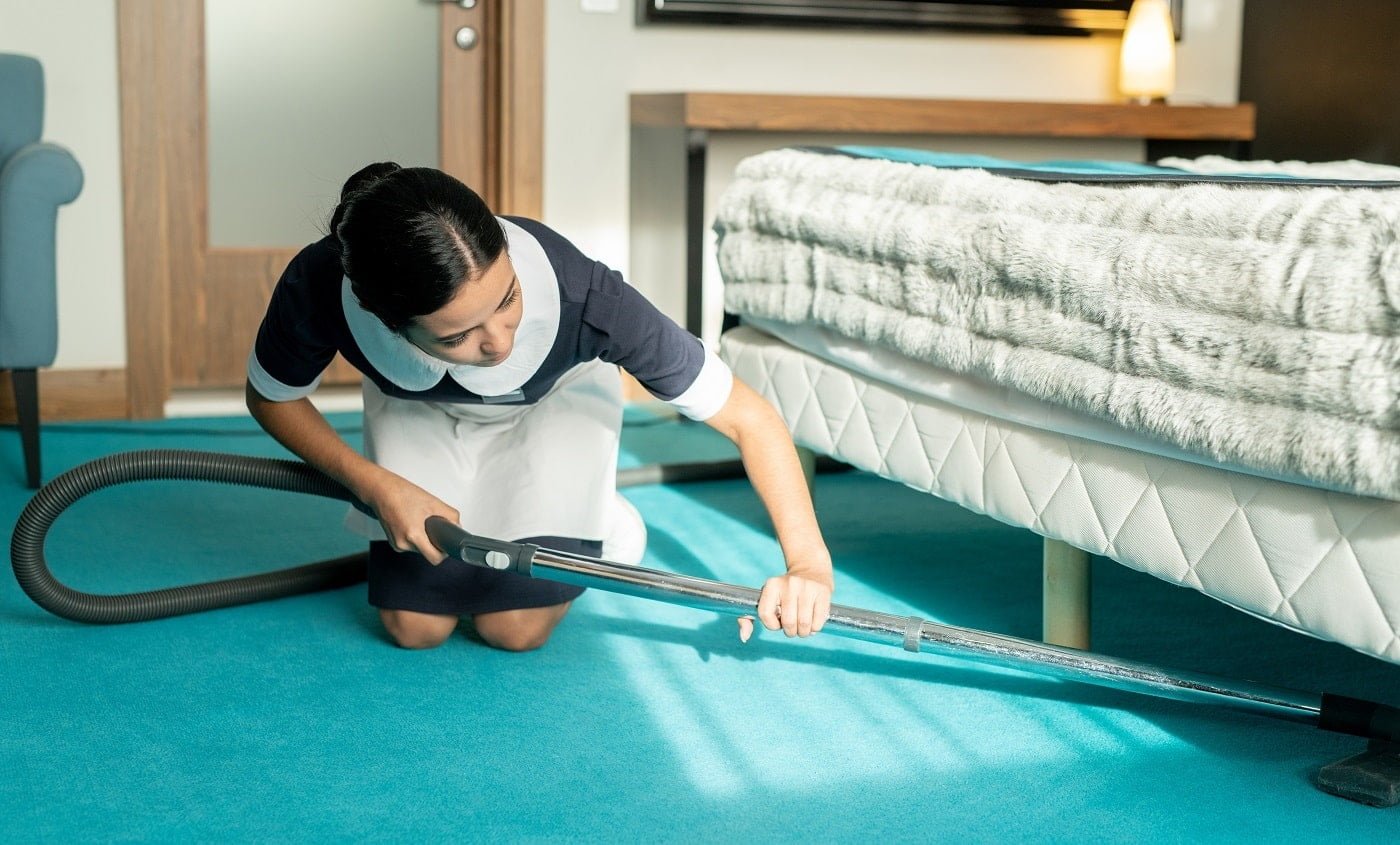 Young beautiful brunette chambermaid in uniform using vacuum cleaner while cleaning blue floor covering under bed in hotel room