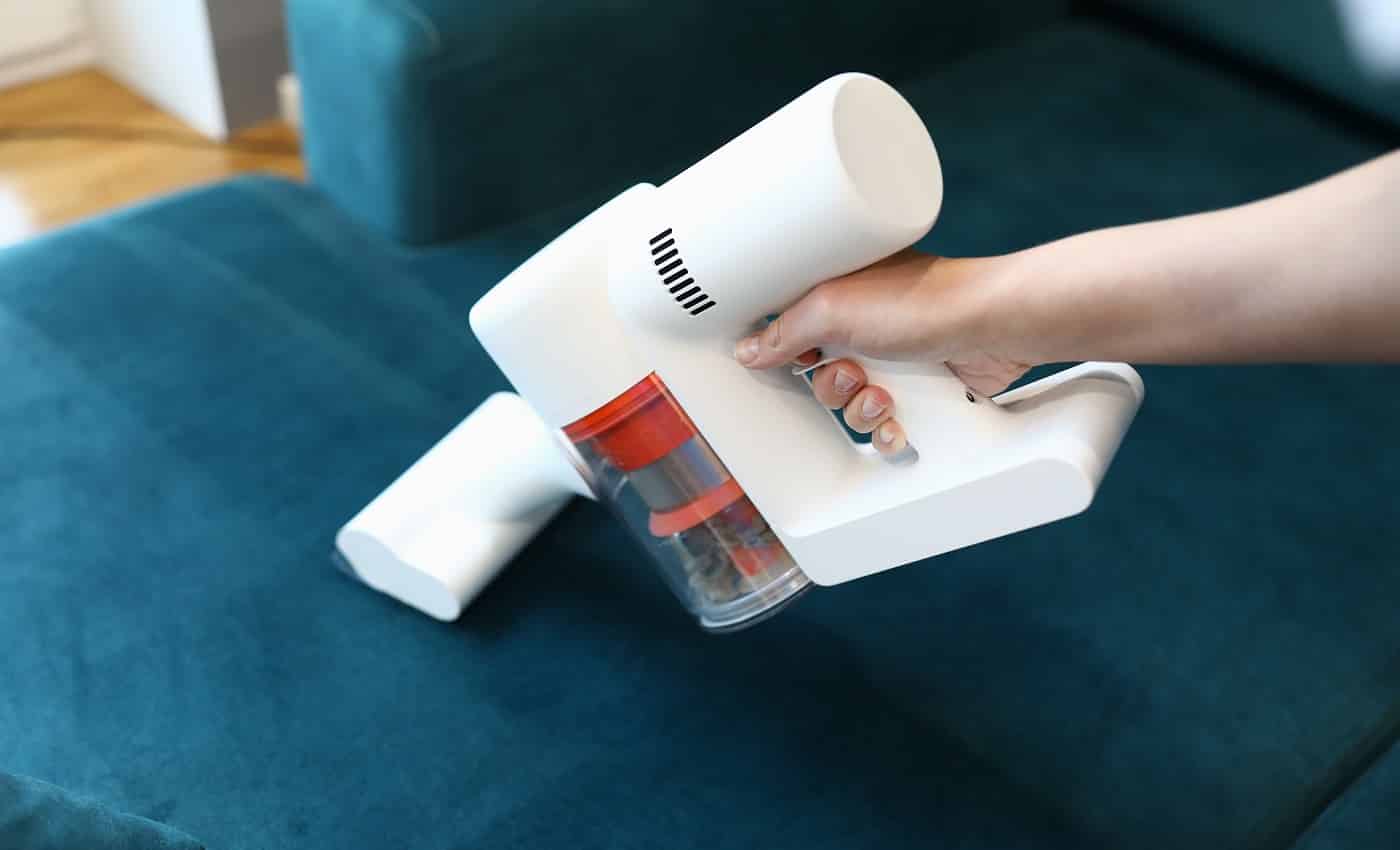 Close-up of womans hand cleaning green sofa with small white compact vacuum cleaner. Renovation and interior design. Housewife or cleaning service concept