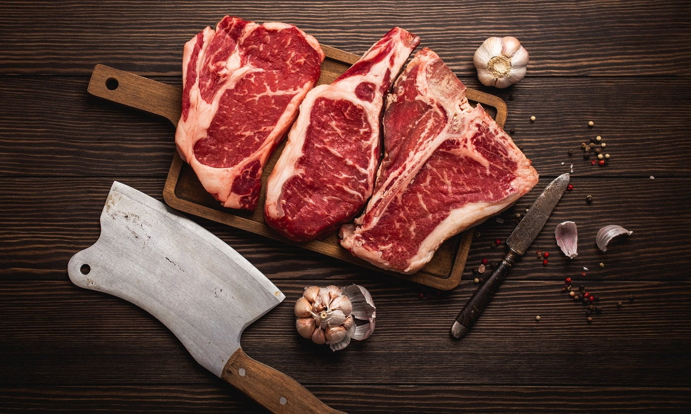 Set of three different types raw meat steaks: Ribeye, T-bone, Cowboy on cut board with knife and seasonings, wooden background. Aged steaks assortment, butchery/restaurant concept, top view, close up