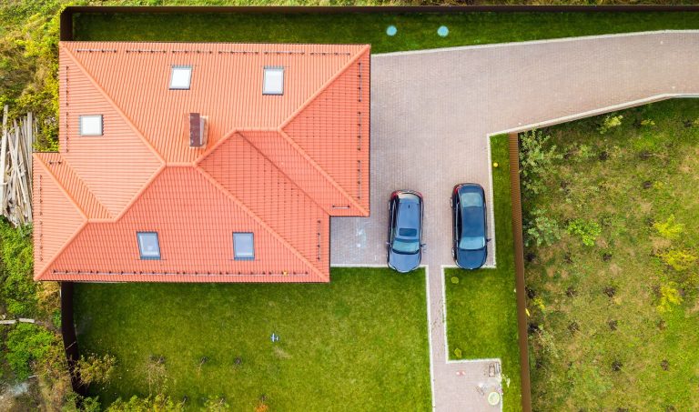 Aerial top view of house shingle roof with attic windows and cars on paved yard with green grass lawn.