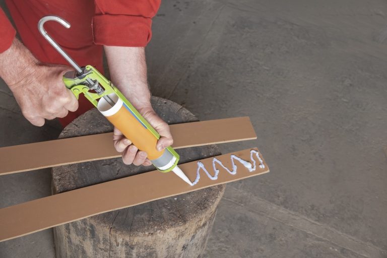 The hands of the worker in a red jumpsuit holding a syringe gun for applying glue. A worker applies white glue to strips of laminated fiberboard, laminate.