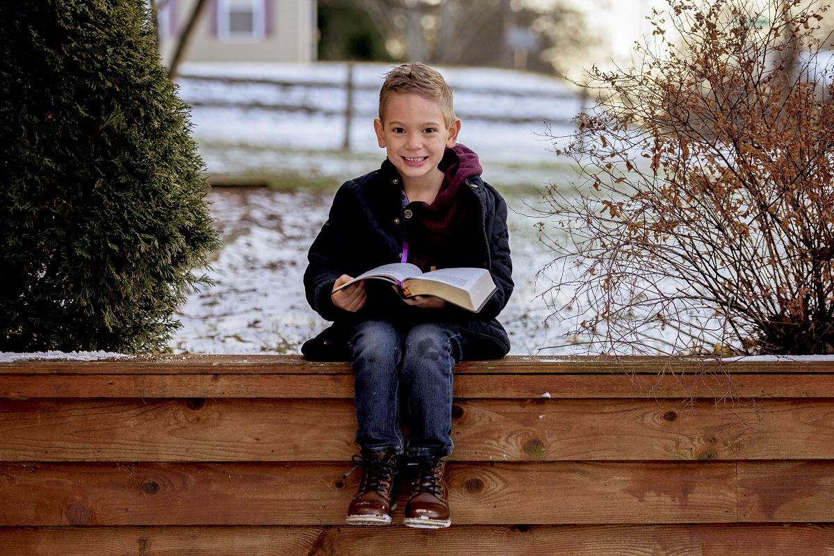 A happy smiling boy sitting on a wooden fence and reading a book in a park