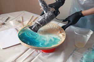 A girl artist in black gloves pours epoxy resin on a round surface and makes an artistic picture of the sea . Master class, hobby.