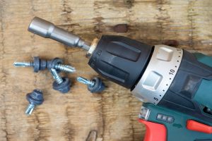 Close-up of a cordless screwdriver lying on a wooden surface with a nozzle for roofing screws. Self-tapping screws painted in gray are lying nearby. Top view, flat lay