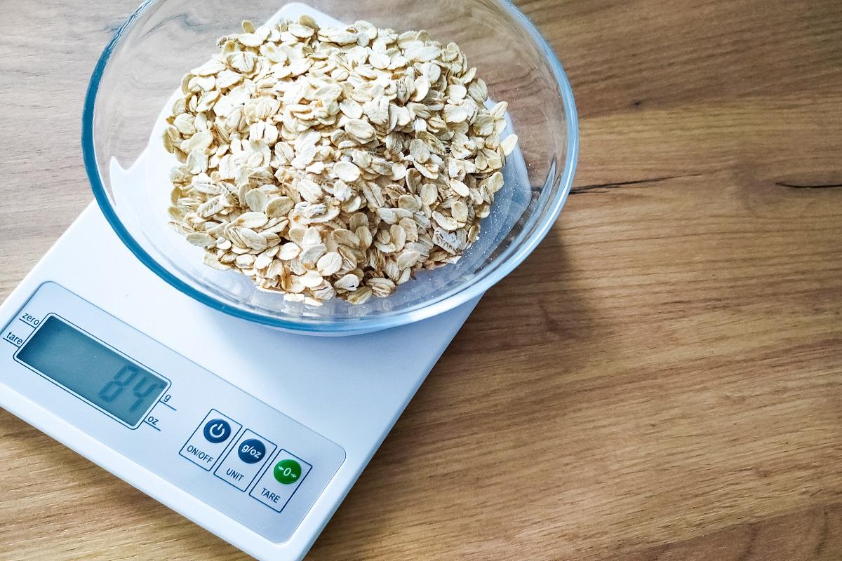 Oat groats, grits weighing in glass bowl on electronic kitchen scales on wooden table.
