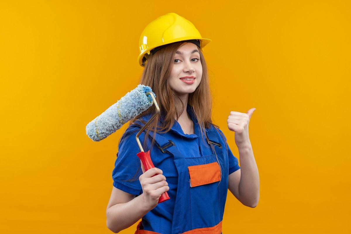 young woman builder worker in construction uniform and safety helmet holding paint roller showing thumbs up smiling confident standing over orange background