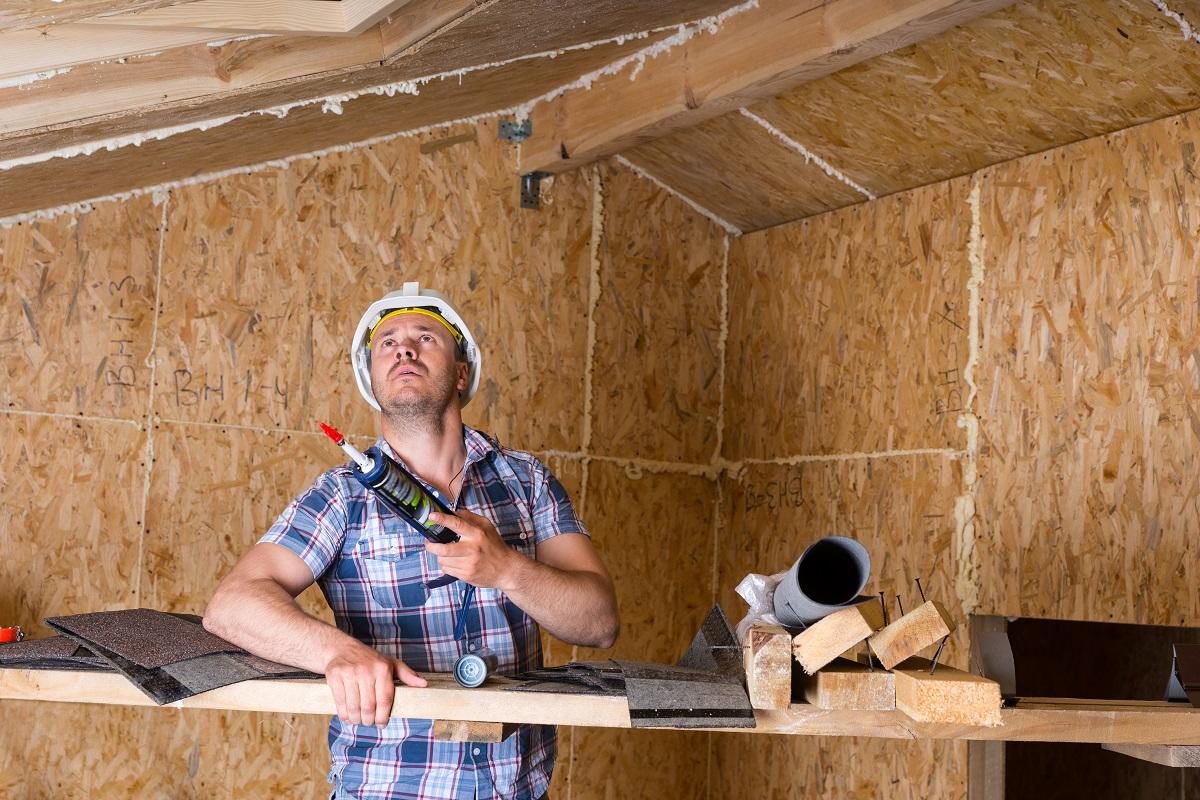 Male Construction Worker Builder Holding Caulking Gun and Looking Up at Ceiling Inside Unfinished Home with Exposed Particle Plywood Board
