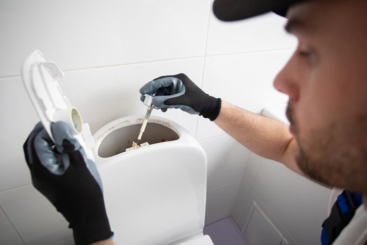 Professional plumber in uniform is repairing toilet bowl in the domestic bathroom with the help of tools