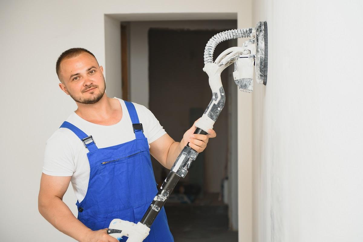 Plasterer smoothes the wall surface with a wall grinder. Master builder grind a white plaster wall. a man in overalls grinds the surface in a respirator. experienced repairman. Best Drywall Sanders.