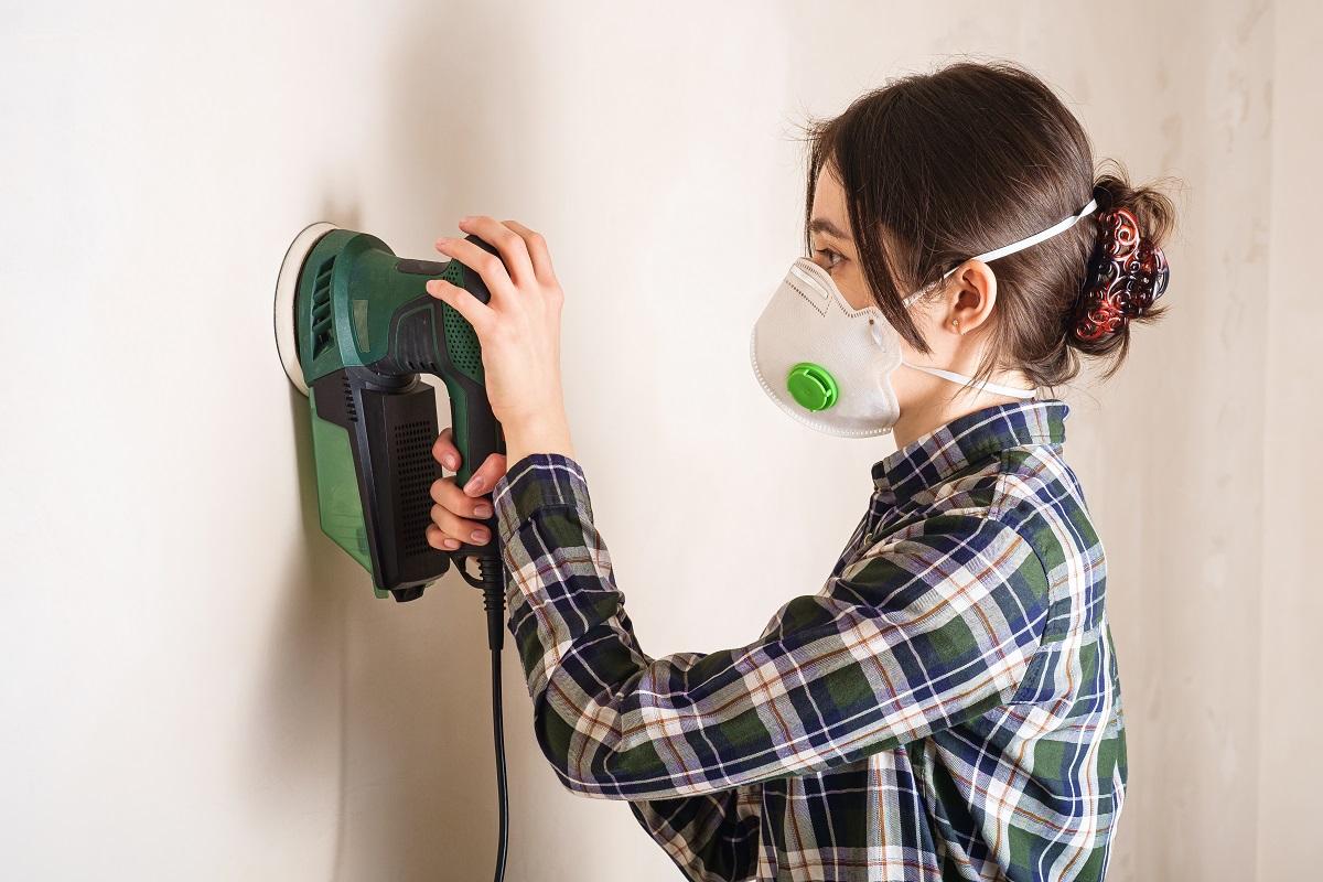 Woman in protective mask working with electric sander to smooth plaster wall surface, room renovation concept. Best Drywall Sanders.