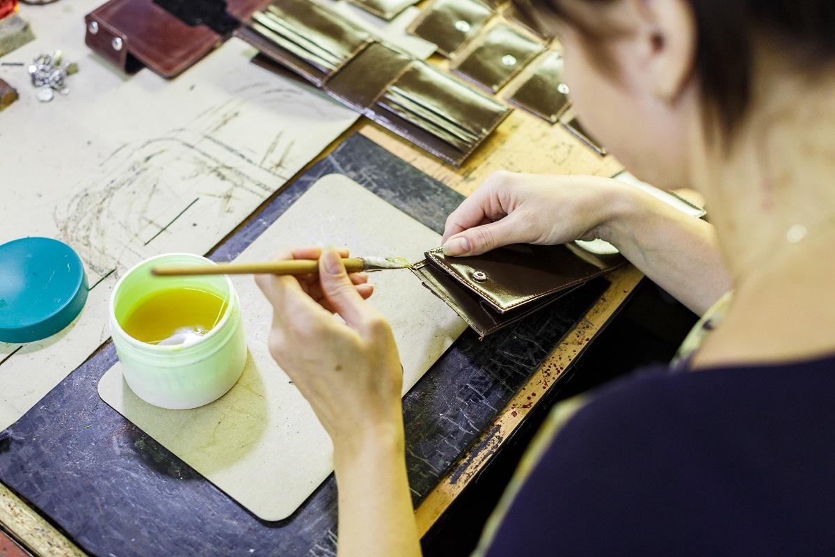 brunette woman smearing with glue a part of a purse with a brush on the wooden table. manufacturing process. close-up photo. How Do You Glue Two Pieces of Leather Together.