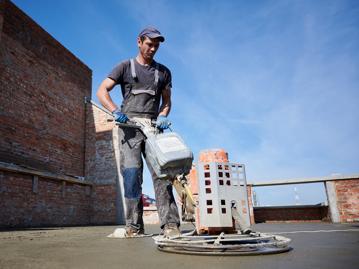 Full length of man laborer using troweling machine while screeding floor in new building under blue sky. Male worker finishing concrete surface with floor screed grinder machine at construction site. Can Old Concrete Be Resurfaced Final Words.