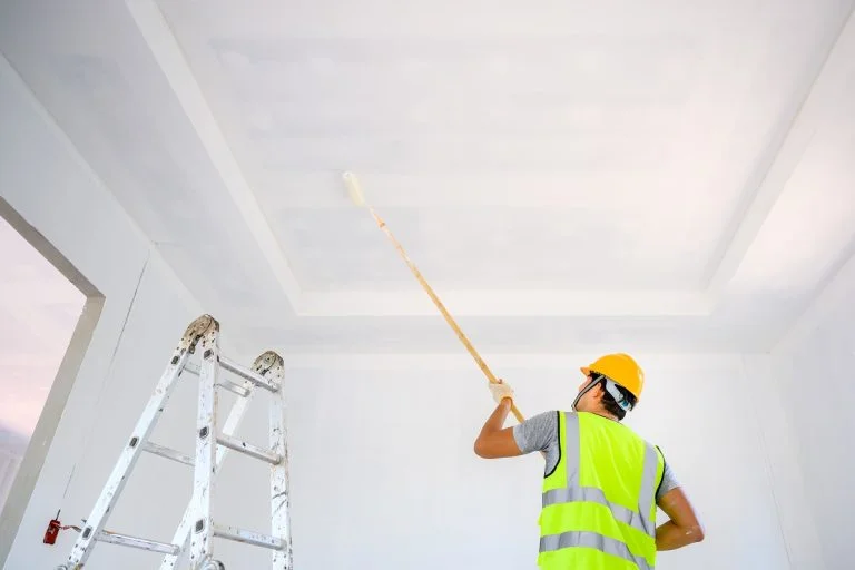 Young Asian male construction worker Work as a house painter Painting the ceiling inside the house and using a white primer paint roller on the construction site. How To Paint A Ceiling With A Roller.