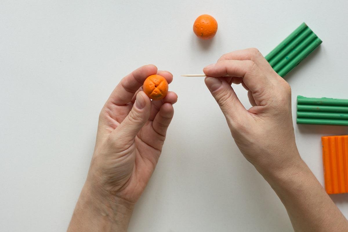 Woman hands showing dot on orange ball made by tooth stick to create orange fruit from the polymer clay. Can You Bake Modeling Clays Of All Types.