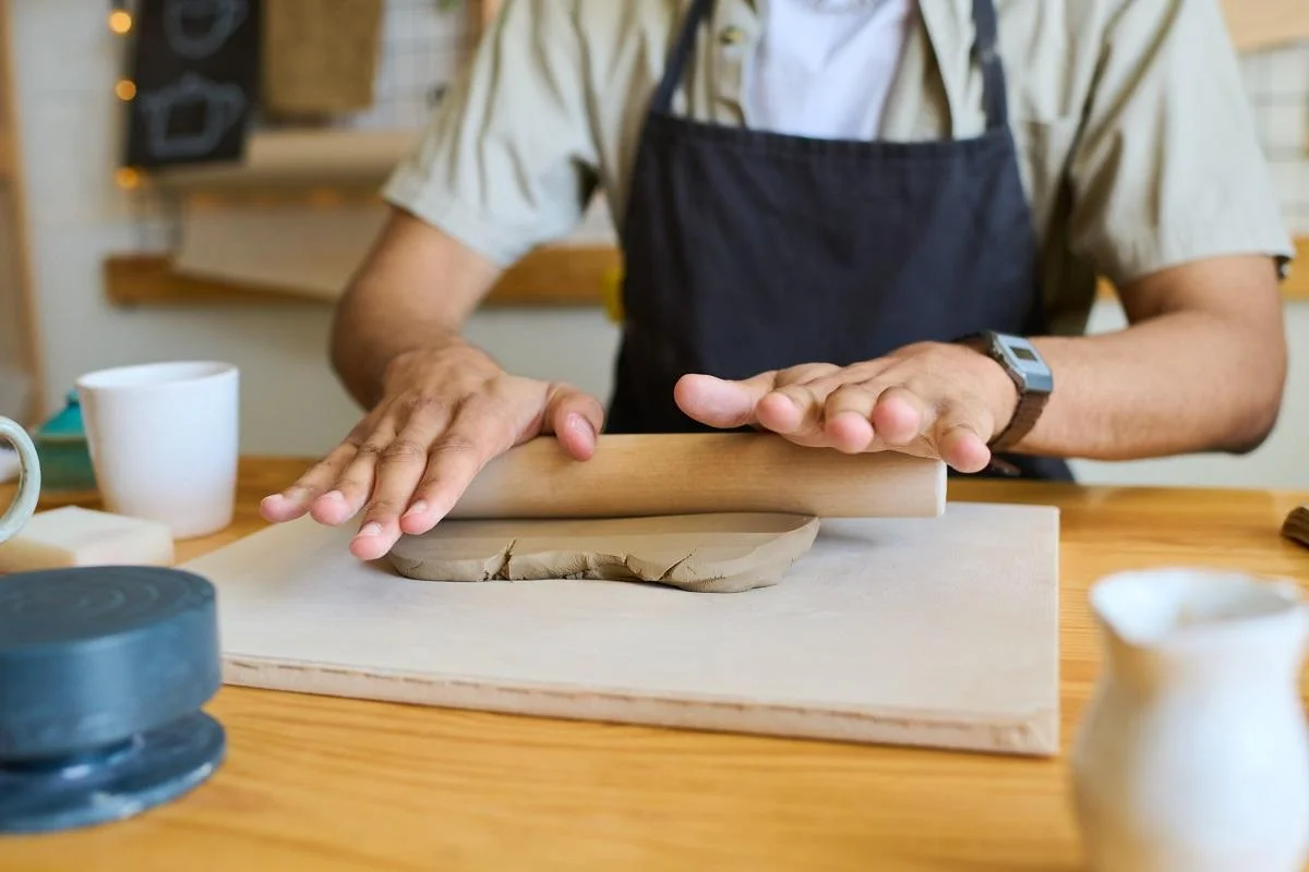 Hands of young black man in apron flattening piece of clay with rolling pin on board while sitting by table with supplies for handwork. The Different Kinds Of Modeling Clay.