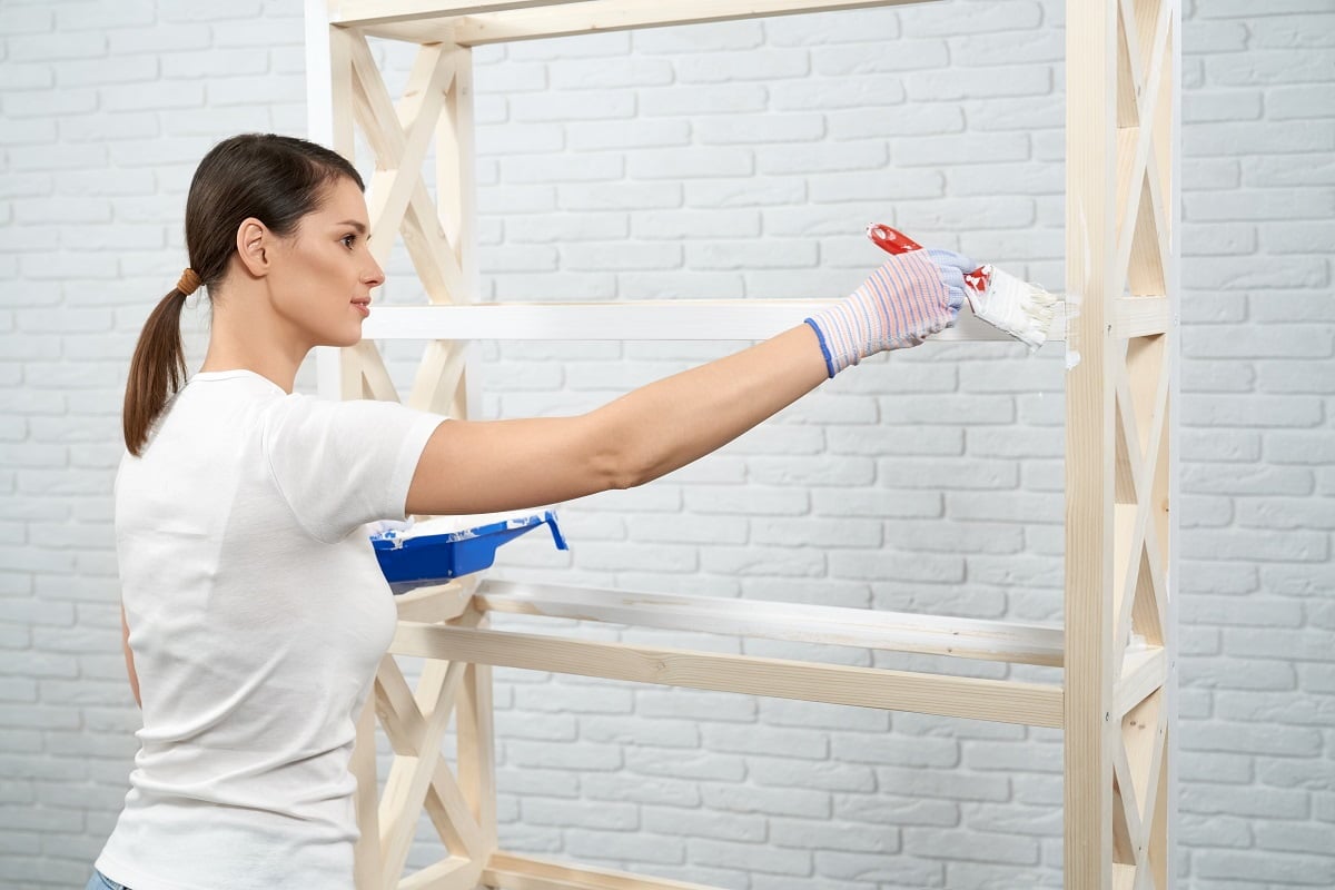 Young woman using brush and white color for painting wooden storage rack indoors. Happy brunette wearing protective gloves during repairing work at home. How To Paint Wooden Furniture.