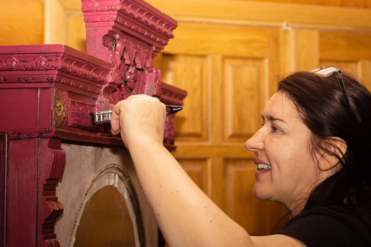 Caucasian woman with smile on face painting wooden ornamented cupboard in red with large paint brush with doors in background. Reuse of old antique things. Home furniture renovation workroom. Paint For Antique Furniture Final Words .