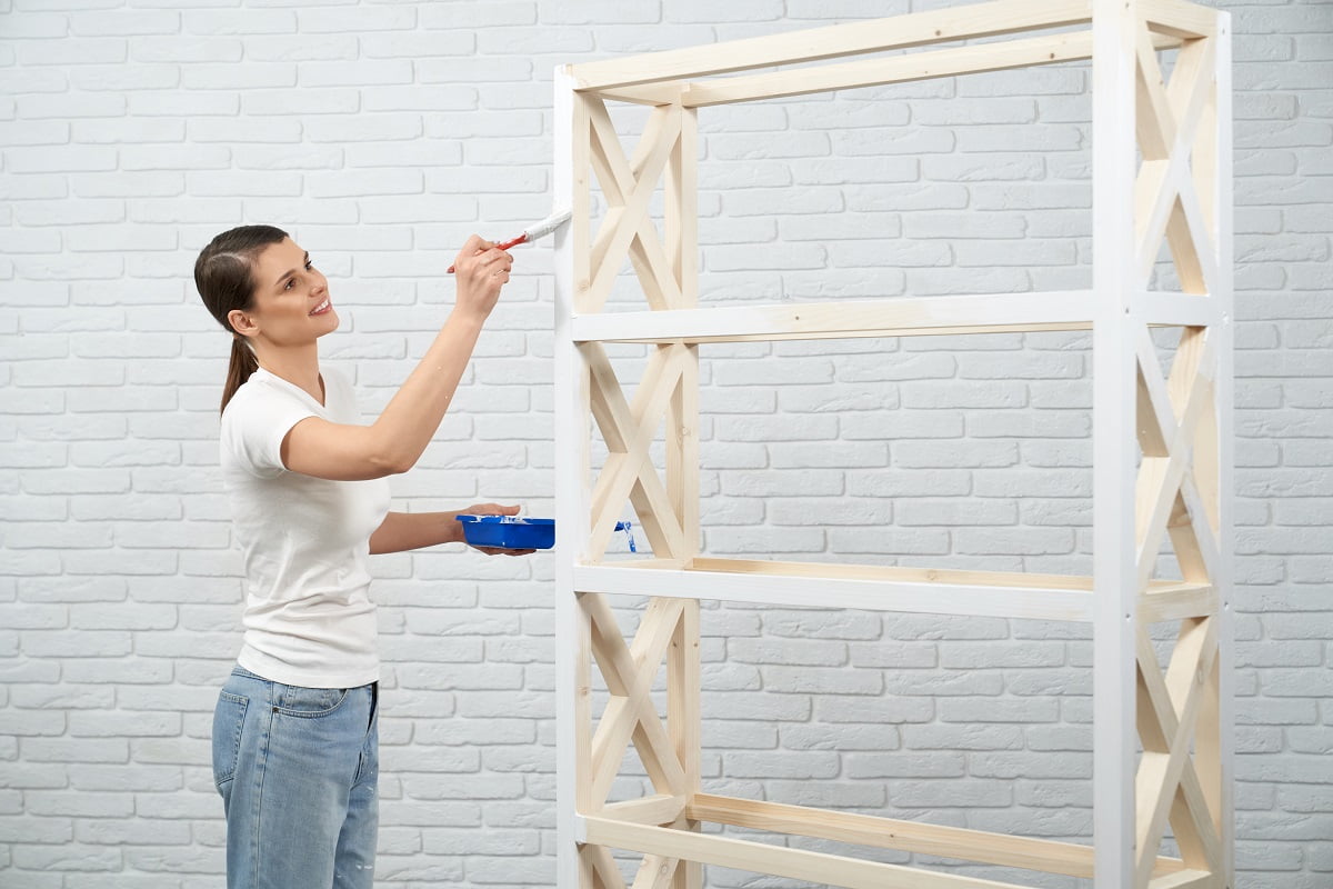 Smiling woman in casual t-shirt and jeans painting in white color wooden shelves. Happy brunette using repair tools for refreshing furniture at home. Preparing To Paint Furniture.