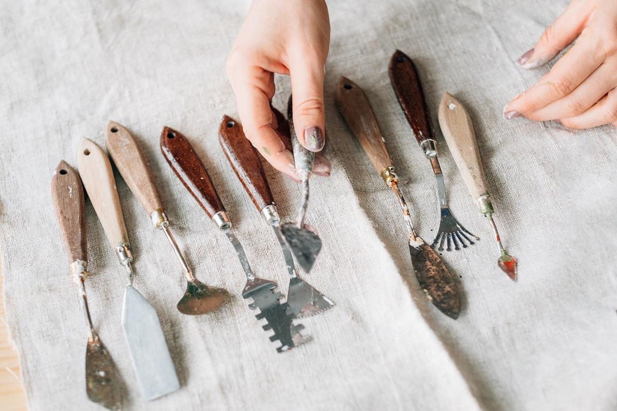 Artist and artwork in process. Craft tools arranged on ivory textile background. Woman hand with palette knife. The Best Clay Sculpting Tools Of The Year.