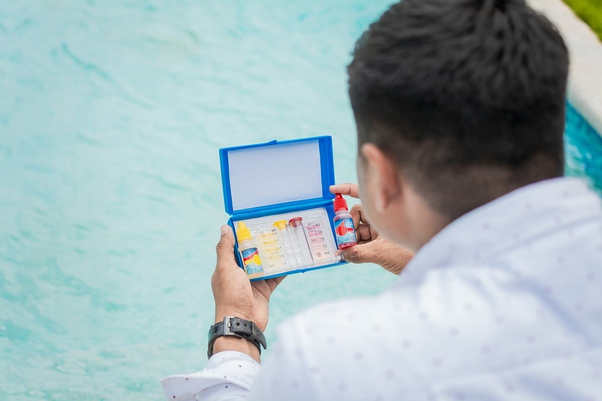 Person holding complete water test kit with blurred pool background, Hand holding Water Test Kit on blurred pool background, Hand holding a pool ph and chlorine tester. Why Do You Need To Test Pool Water.