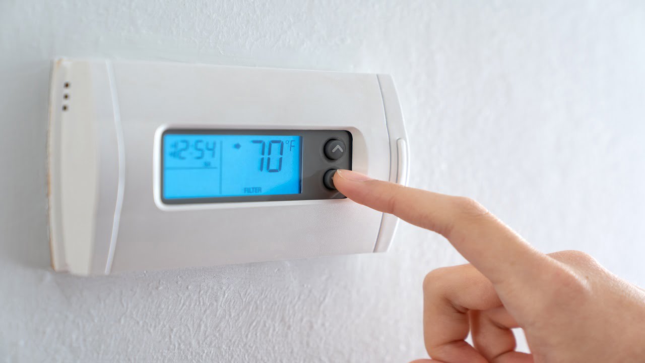 A woman is pressing the down button of a wall attached house thermostat with digital display showing temperature 70 degree Fahrenheit for heating, cooling, electricity and gas saving. Thermostat And Room Temperature.